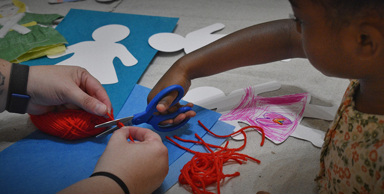 A child cuts yarn held by adult hands to decorate a paper doll restging on a table..