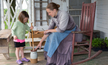 A girl churns butter while woman in a long dress assists her.