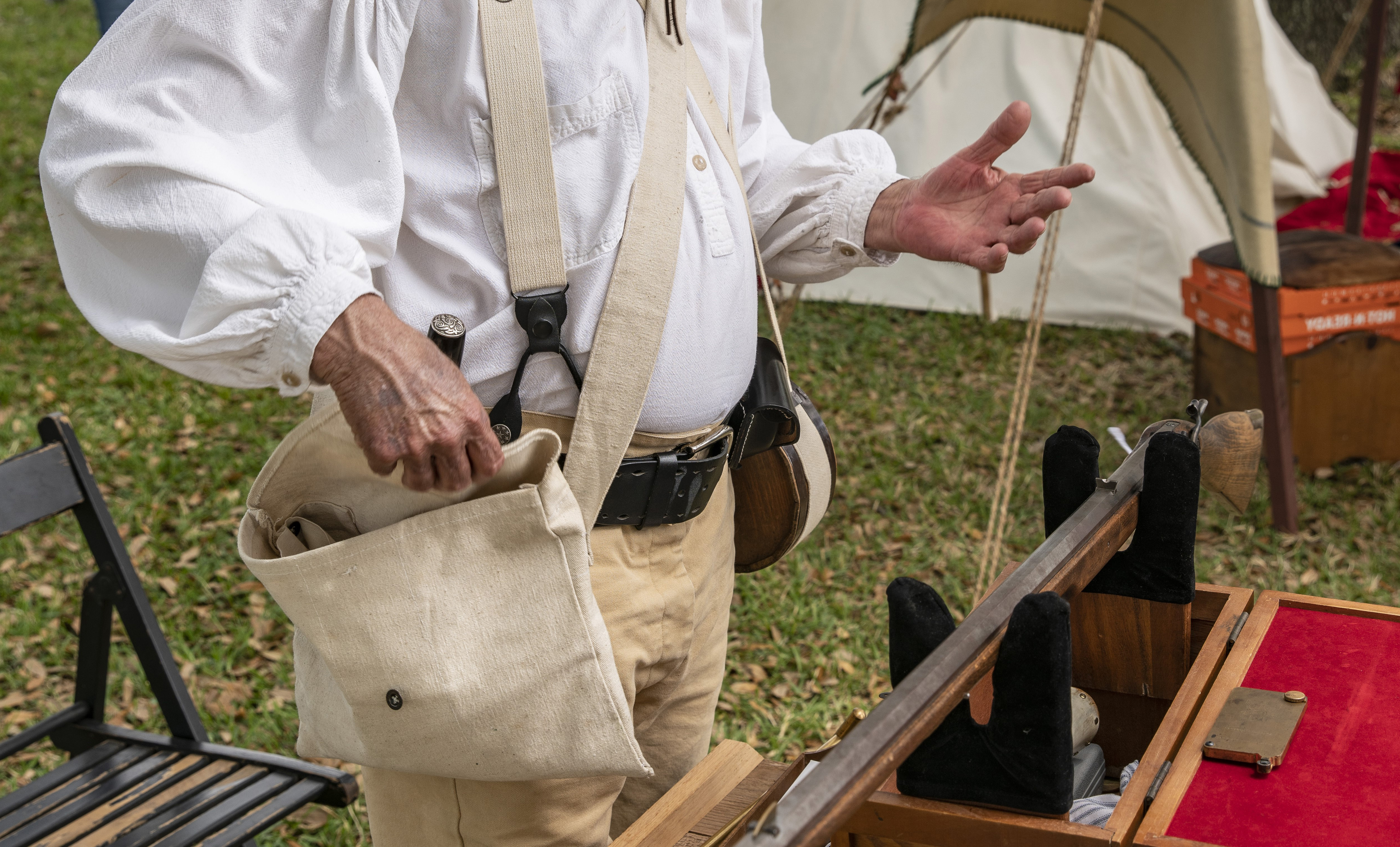 A reenactor opens a canvas bag hanging from his shoulder while gesturing with his other hand.