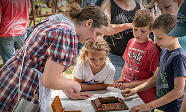A woman wearing a plaid dress leans over a table showing items to children.