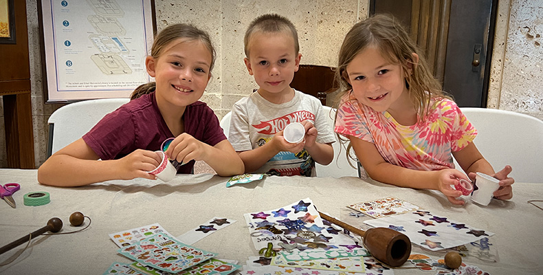 Three smiling children at a table making a cup and ball toy