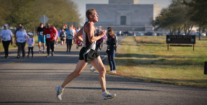A man wearing athletic clothing runs from left to right; behind him dozens of people walk from the San Jacinto Monuemtn