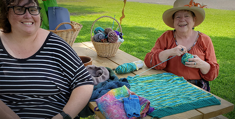 Two woem sitting outside at a wooden picnic table smiling and knitting with colorful yarn.