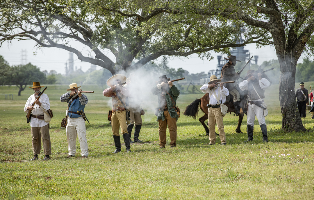 Reenactors fire muskets while standing in a line under a tree.