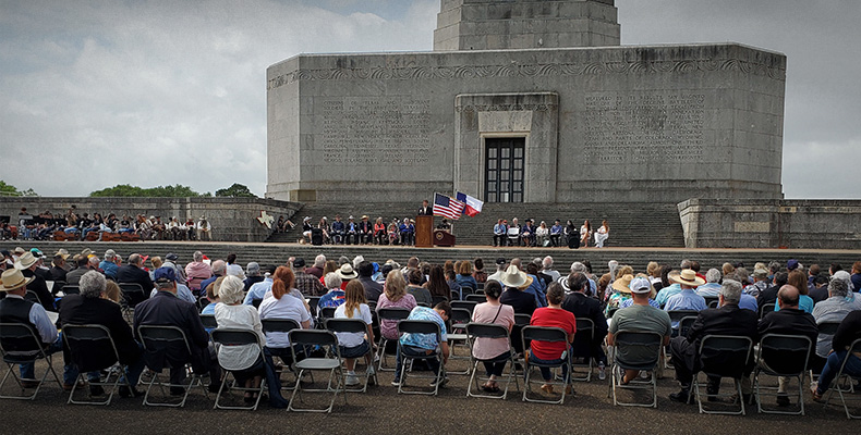 People sit in rows of chairs with their backs to the camera, facing a tall stone building and other people seated on a raised terrace, with Texas and U.S. flags flying.