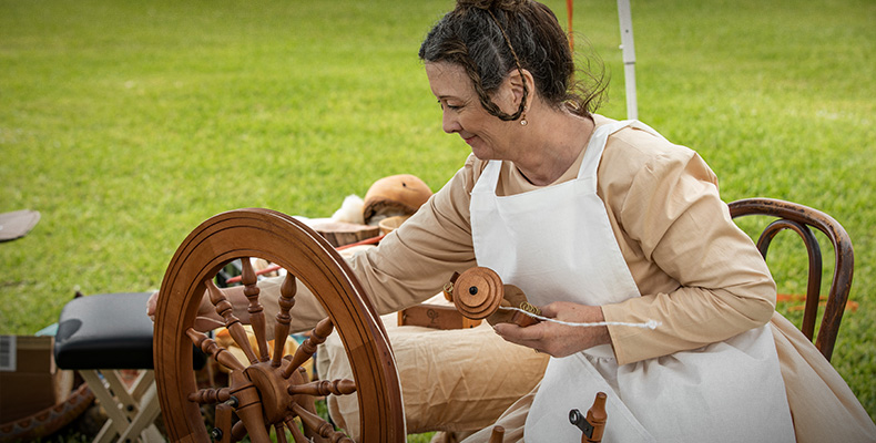 A woman sits at a spinning wheel, with a young child sitting on the floor near by.  Text reads: Life on the Frontier: Spinning and Weaving