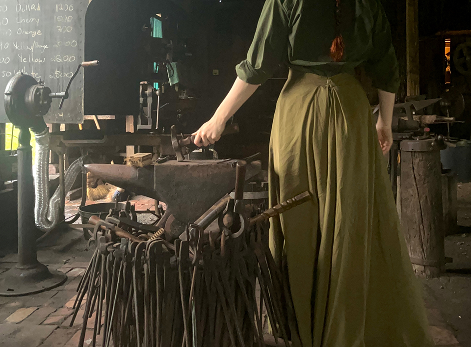 A woman holds a blacksmithing tool on an anvil, surrounded by other blacksmithing equipment.