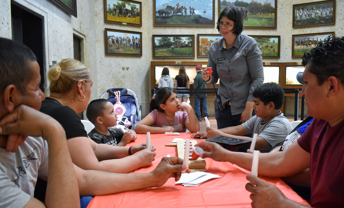 A woman holding a cup demonstrates a skill to a group of seated children and adults.