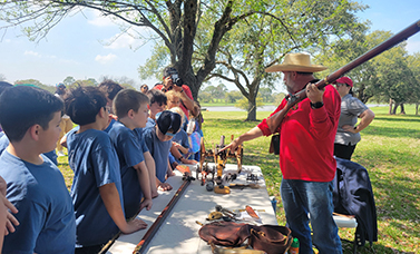 A man holding a replica firearm explains the gun to children standing on the other side of a table.