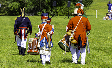 Drummers and fifers in red military uniforms march away from the viewer, following an officer in a blue coat.