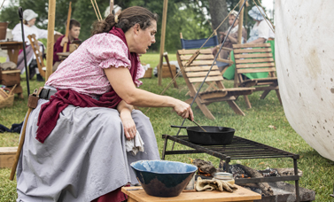 A woman sitting by a fire stirs food in an iron skillet over a camp fire.