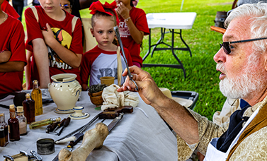 A man holds up a medical tool, with medical equipment on a table viewed by children.