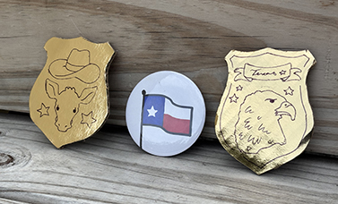 Two gold-colored badges with symbols drawn on in black; and a button with a Texas flag. showing an exploding bomb.