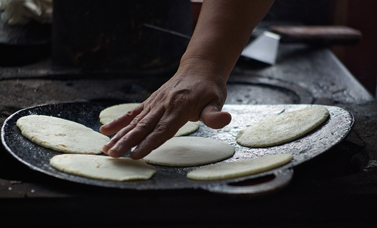 A hand pats out a tortilla on a metal griddle.