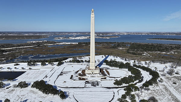 An aerial view of the San Jacinto Monument surrounded by snow, under a blue sky.