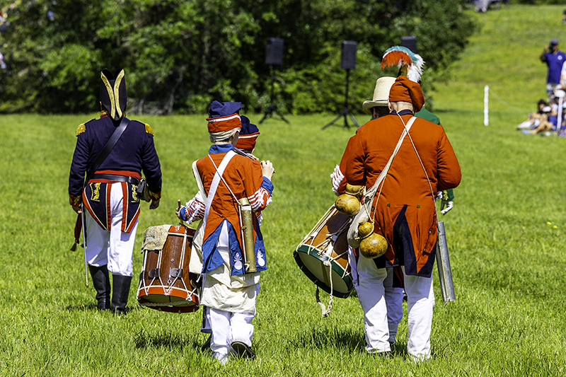 Drummers and fifers in red and blue uniforms march away from the viewer, following an officer in a blue coat.