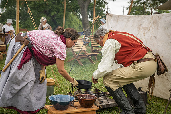A woman holding an iron skillet, and a man, stoop over a cooking fire.