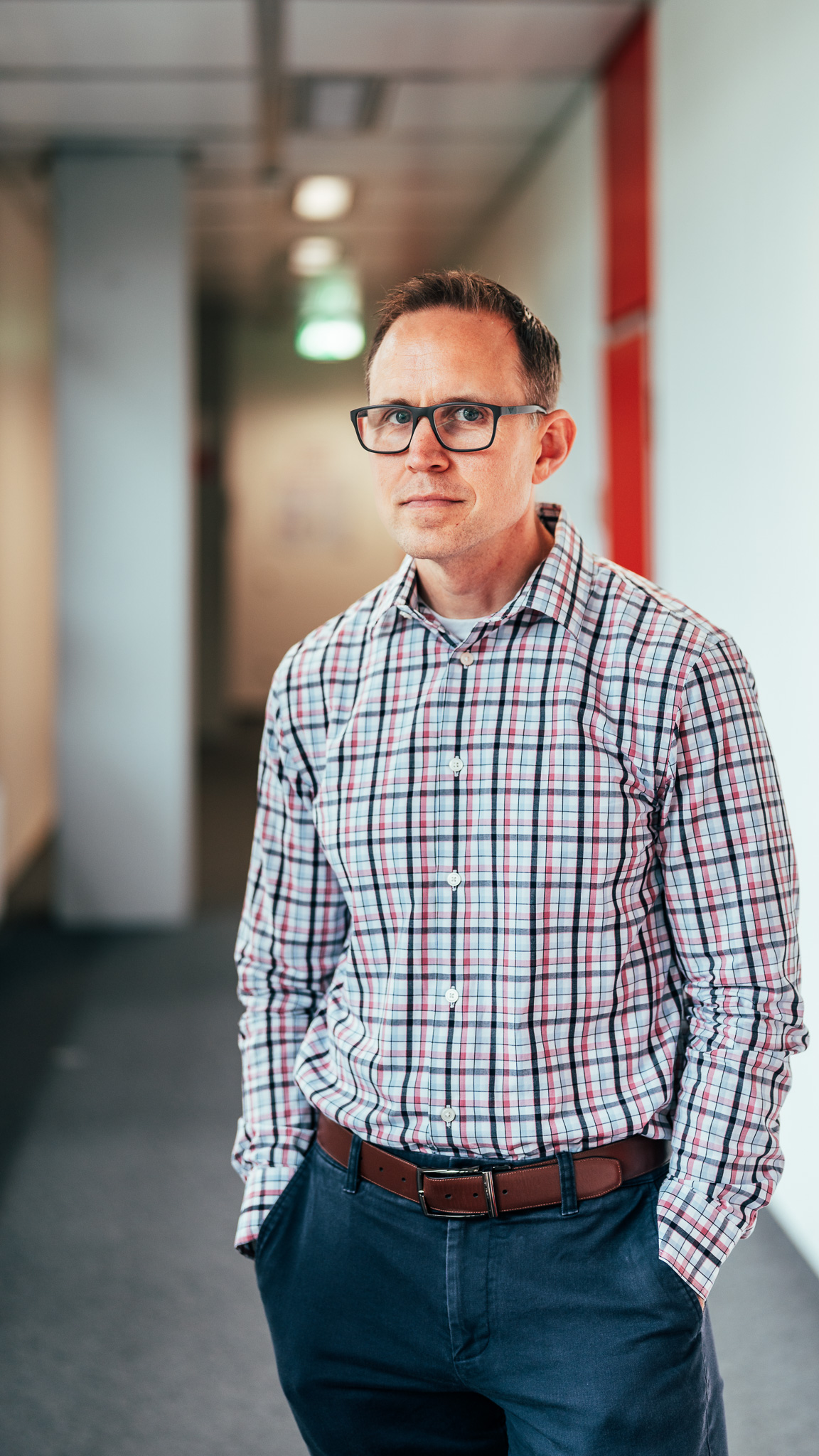 A man wearing a plaid shirt stands in a hallway looking at the camera.