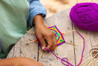 Hands are weaving yarn around crossed sticks.