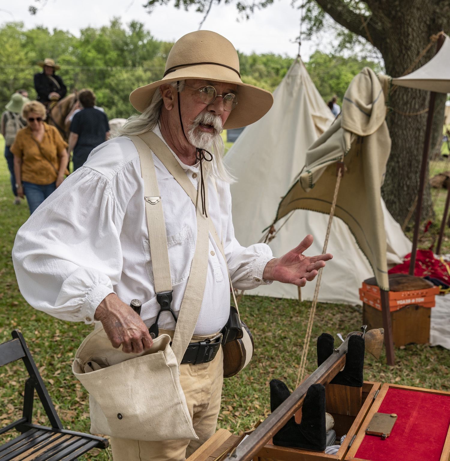 A reenactor reaches into a canvas bag hanging over his shoulder while gesturing with his other hand.