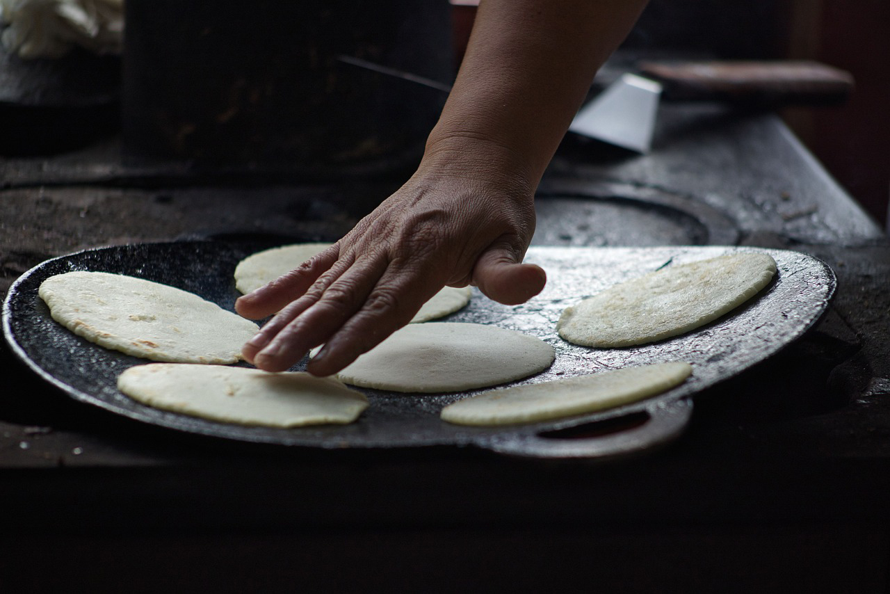 A hand pats out a tortilla on a metal griddle.