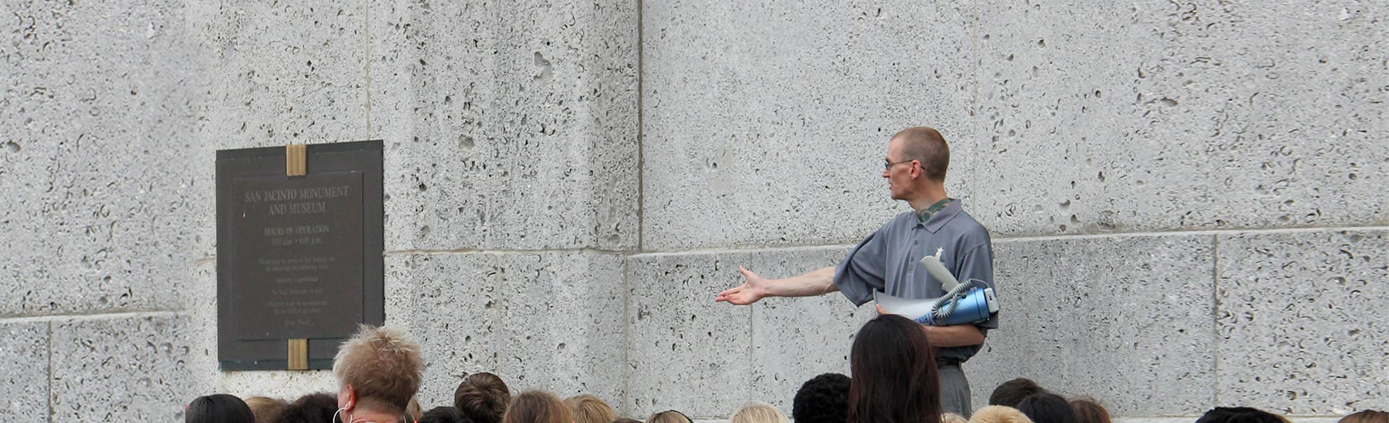 A tour guide gesturing to a plaque on the outside wall of the San Jacinto Monument