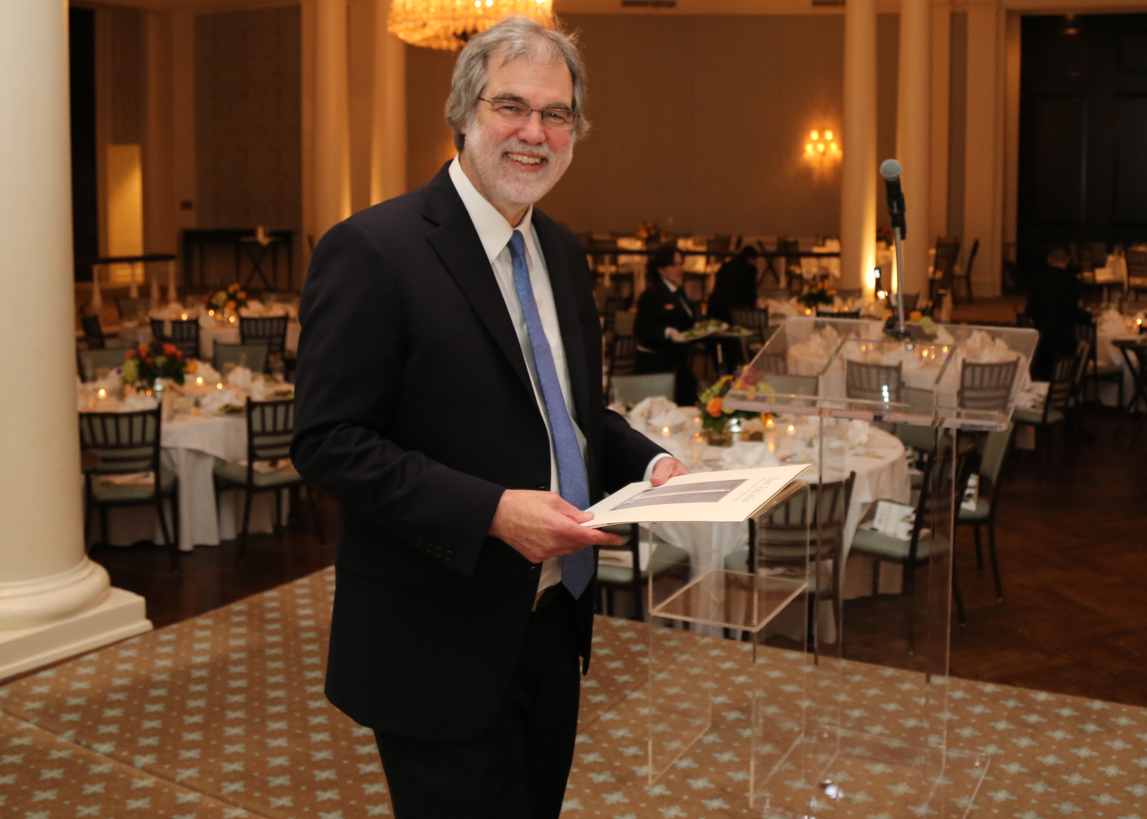 A man in a dark suit smiles at the camera; behind him are tables set for dinner.