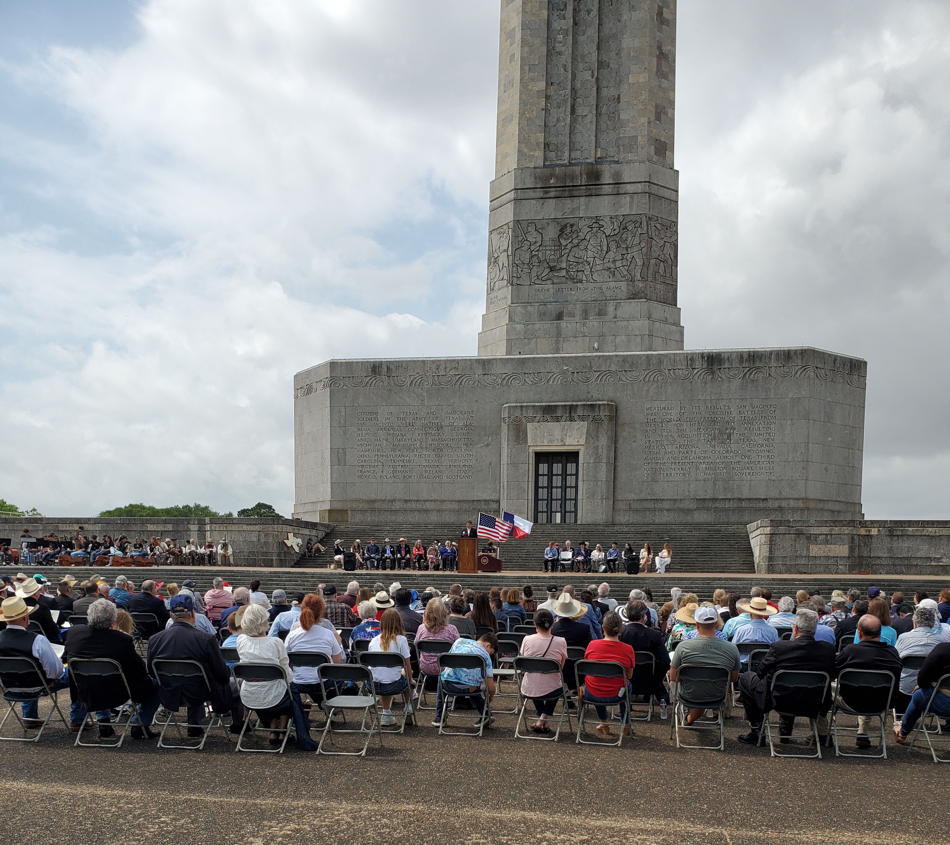 People sit in rows of chairs with their backs to the camera, facing a tall stone building and other people seated on a raised terrace, with Texas and U.S. flags flying.