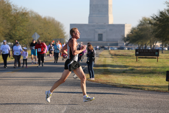 A man wearing athletic clothing runs from left to right; behind him dozens of people walk from the San Jacinto Monuemtn