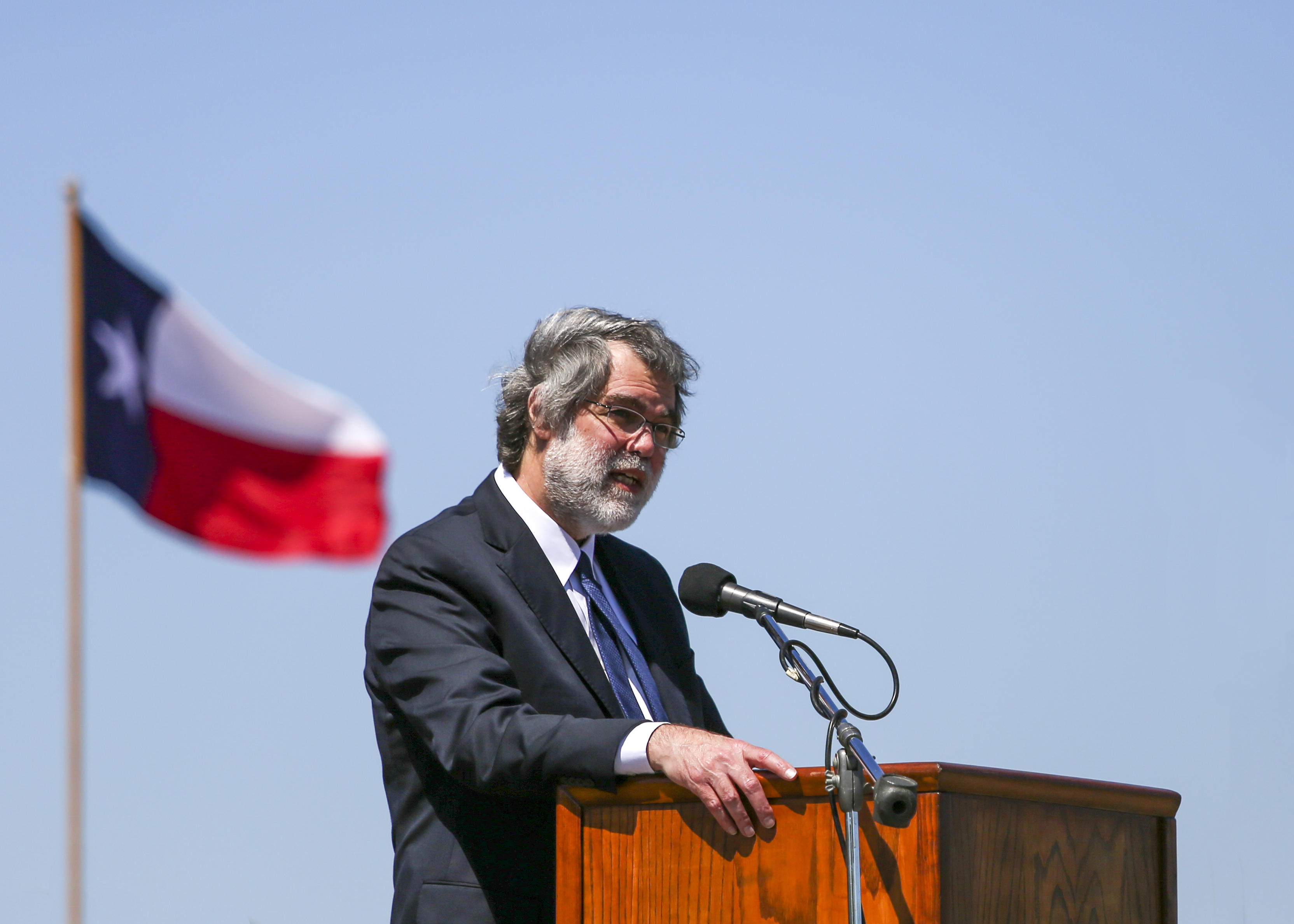 A man wearing a suit stands at a podium outside, with a Texas flag behind him.