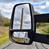 Picture looking at a rearview mirror on a vehicle that shows the tall obelisk of the San Jacinto Monument, with trees in the background.
